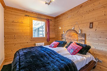Cozy chalet bedroom with warm wood paneling, a bed draped in a dark furry throw, plaid cushions, and a unique circular headboard.