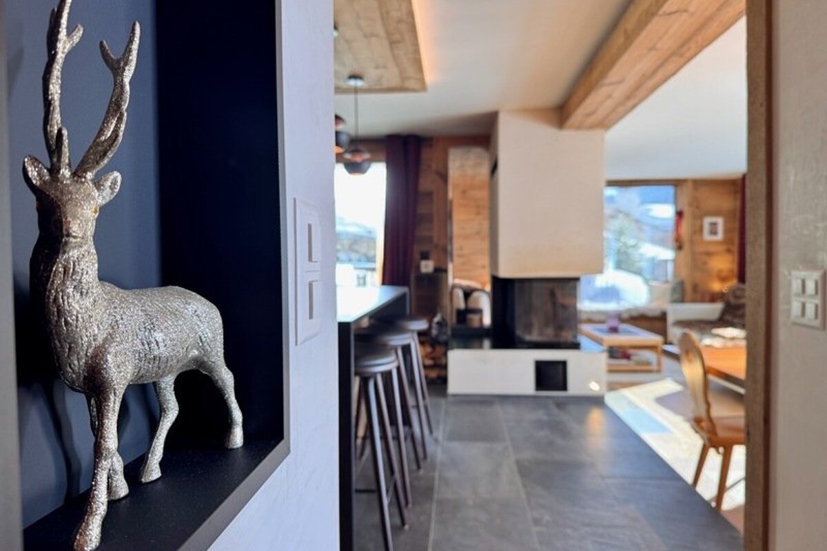 A silver stag figurine in a wall niche overlooks a modern, warm chalet interior with a fireplace in the background.