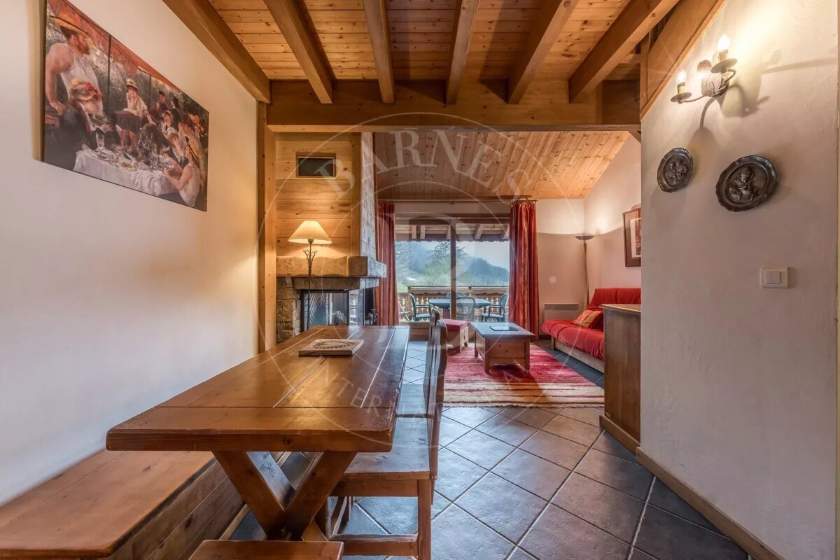 Cozy chalet interior with a rustic wooden dining table, stone fireplace, red sofa, exposed beams, and a balcony view of mountains.