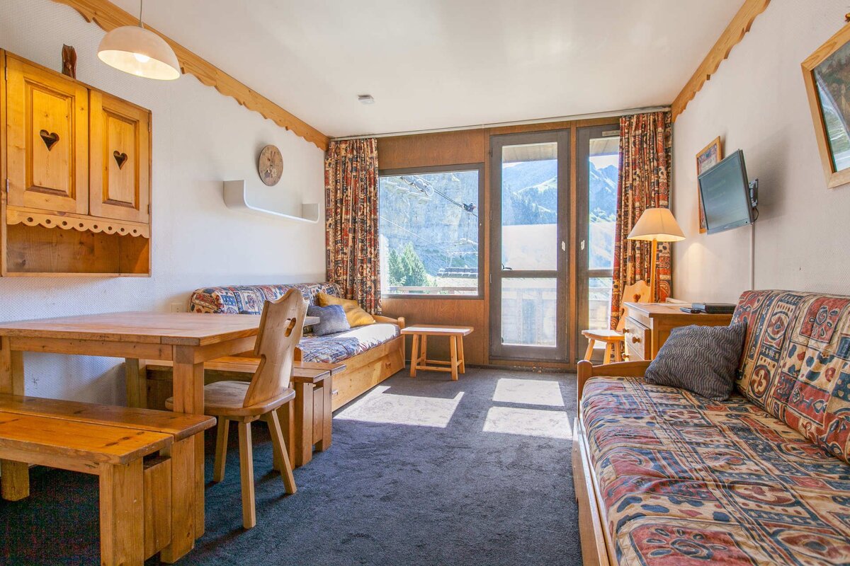 A cozy, rustic living/dining room with wooden furniture, patterned upholstery, a wall-mounted TV, and large windows overlooking a mountain landscape.