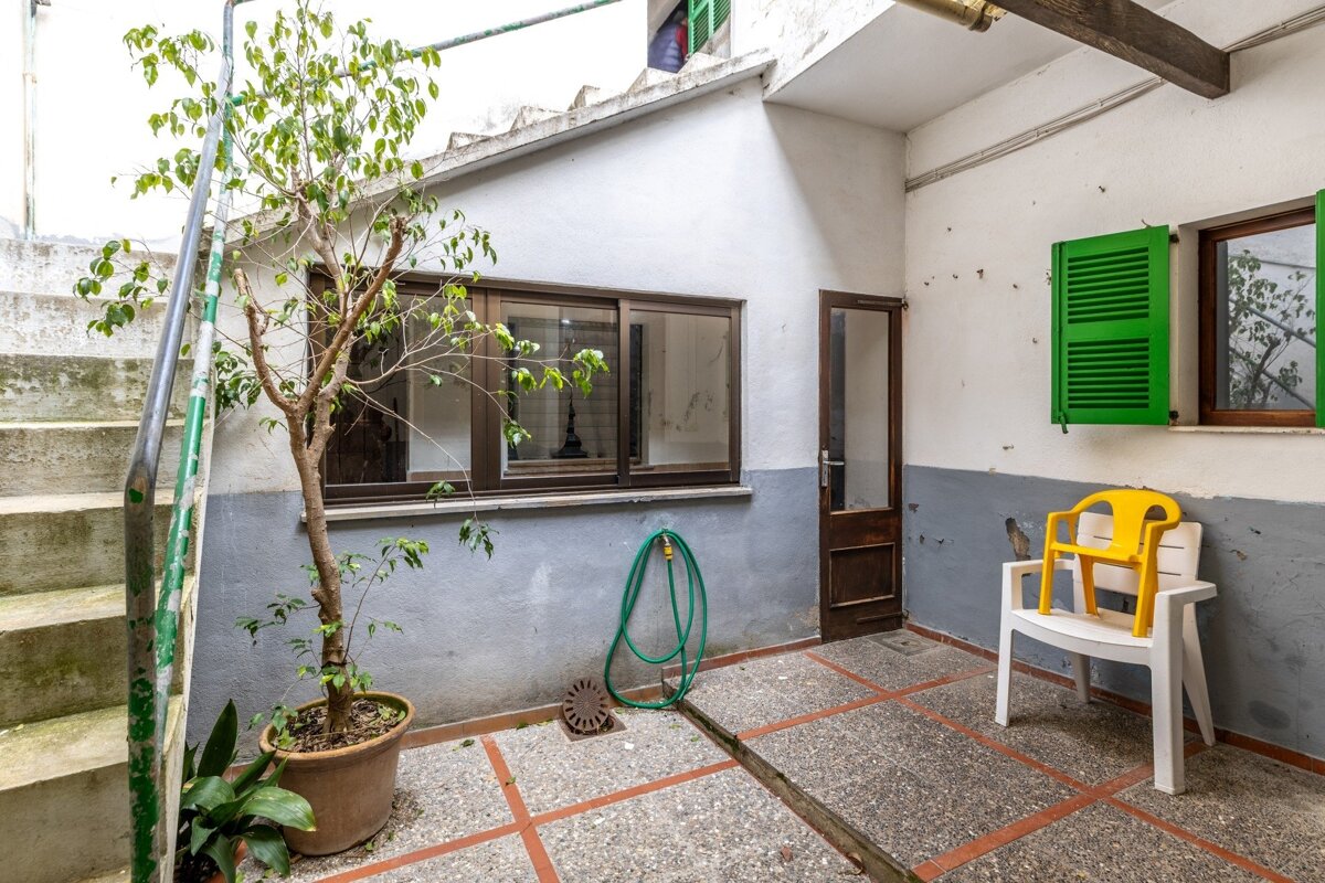 A small, worn outdoor patio with a potted tree, concrete stairs, windows with green shutters, a garden hose, and plastic chairs on a tiled floor.
