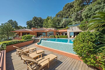 Luxurious terracotta villa featuring a curved pool, wooden deck with sun loungers, and lush green trees under a clear blue sky.