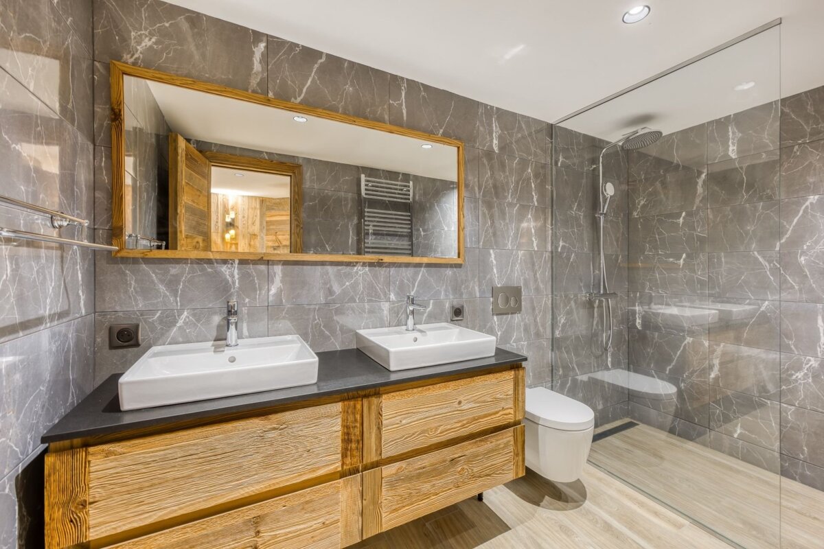 A modern bathroom featuring gray marble-look tiles, a double wooden vanity with white sinks, a large mirror, and a glass shower enclosure.