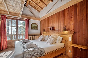 A cozy, rustic bedroom with wooden walls and ceiling, a large bed with bathrobes, and a window revealing a snowy, forested view.