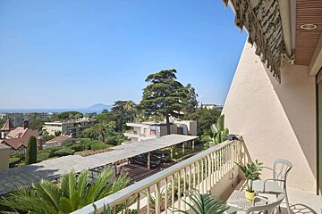 Sunny balcony overlooking a vibrant coastal city with lush trees, buildings, and the blue sea in the distance under a clear sky.