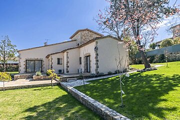 A beautiful stucco house with stone trim and a tiled roof, set in a sunny, manicured garden with a pink flowering tree and stone path.