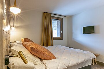 Cozy bedroom with a large bed, white duvet, orange pillows, and brass wall lights. Features a wooden-framed window with a curtain and a wall-mounted TV.