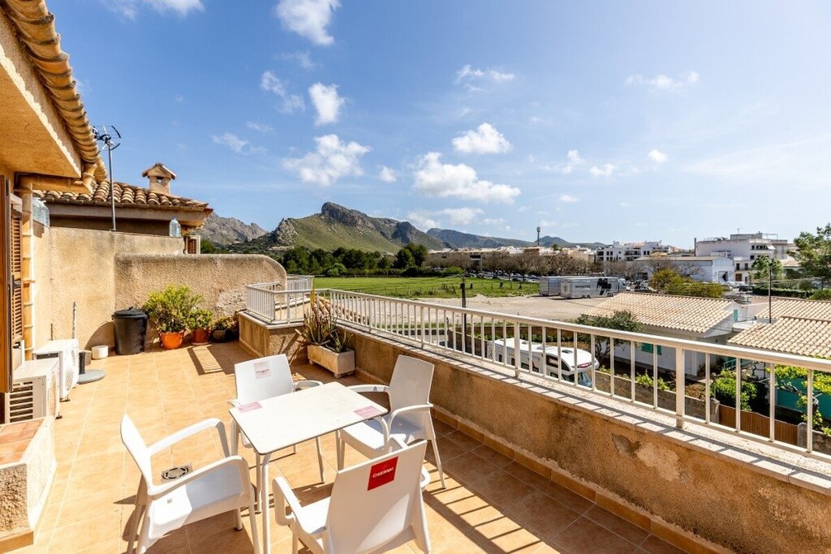 Tiled balcony with a white table and chairs overlooking a town, green fields, and mountains under a bright blue sky.