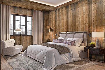 Cozy cabin-style bedroom with wood paneling, a large bed, fluffy white armchair, patterned rug, and warm, inviting lighting.