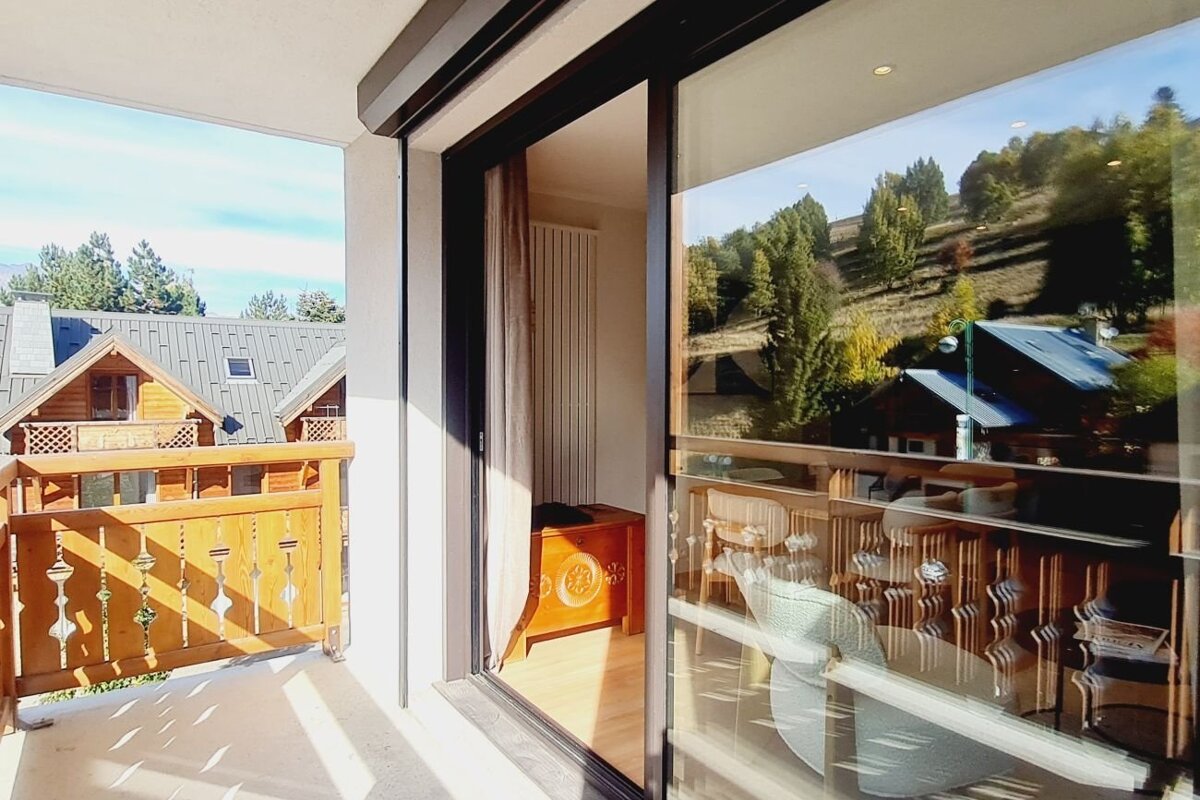Sunny balcony of a chalet with sliding glass doors reflecting a scenic hillside and nearby wooden cabins.