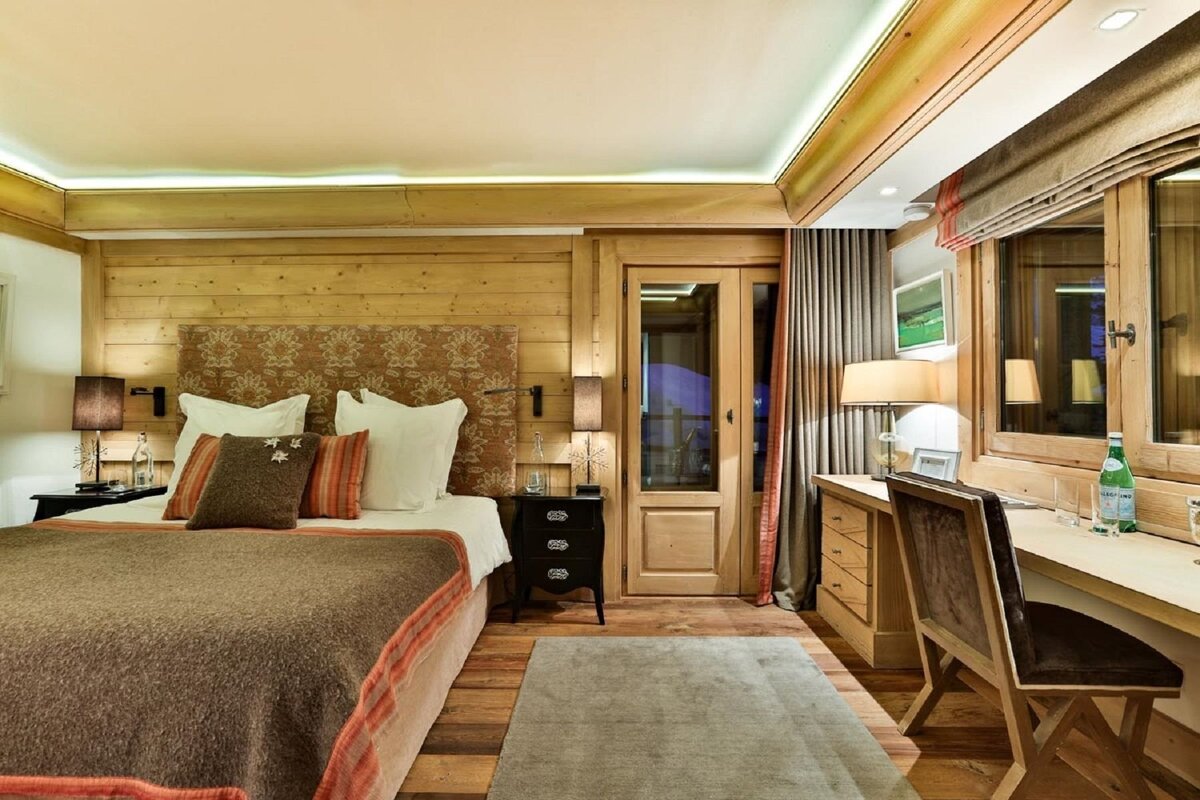 A cozy, rustic bedroom with wood-paneled walls, a large bed, a desk near the window, and a glass door. Warm lighting sets an inviting mood.