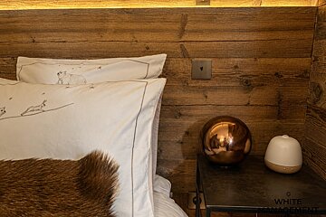 Cozy bed with animal-print pillows against a wood wall, next to a nightstand with a reflective bronze orb and a white diffuser.