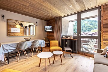 Cozy chalet-style living/dining area with wood accents, a long dining table, modern furniture, and large windows overlooking a scenic mountain valley.