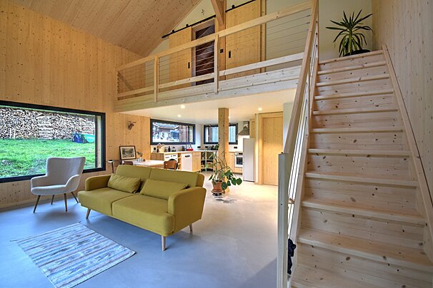 Bright, modern cabin interior with wooden walls, yellow sofa, and open-plan kitchen. Features a loft, wooden staircase, and large window overlooking a stacked woodpile.