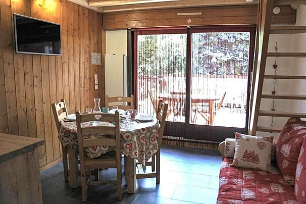 Cozy cabin interior featuring a wooden dining set, a wall-mounted TV, red sofa, and glass doors opening to a sunlit patio.