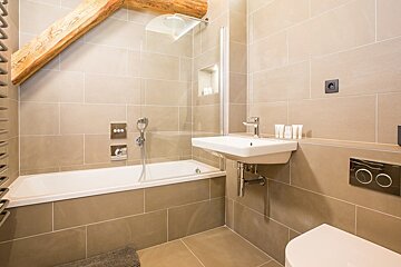 Modern bathroom featuring beige tiles, a bathtub with a glass screen, a sink, and a rustic wooden ceiling beam.