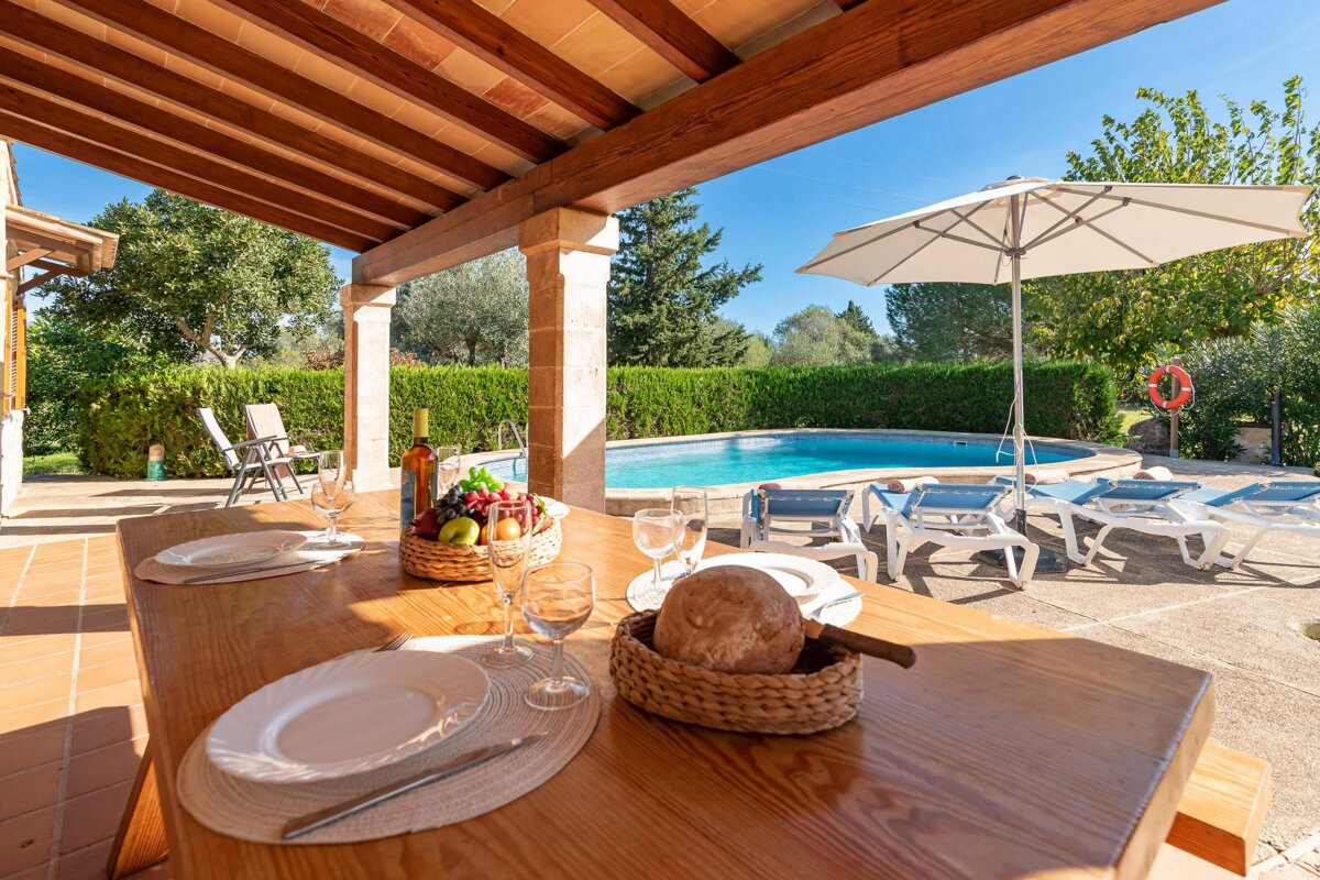 A sunny outdoor patio with a set dining table, fruit, and bread, overlooking a clear blue swimming pool with lounge chairs.