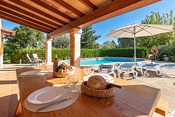 A sunny outdoor patio with a set dining table, fruit, and bread, overlooking a clear blue swimming pool with lounge chairs.
