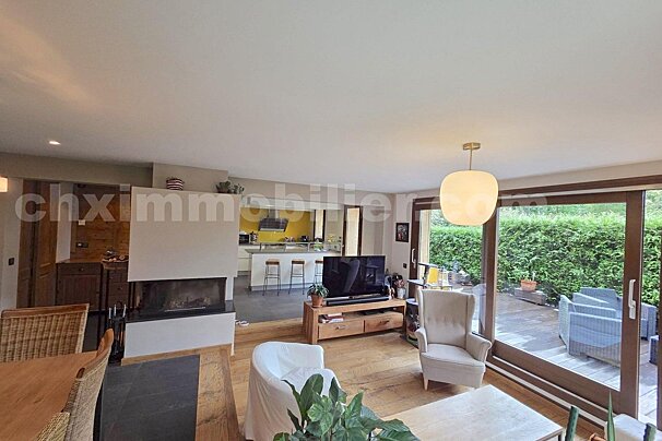 Spacious open-plan living room with a fireplace, TV area, and kitchen, leading to an outdoor patio with garden views.