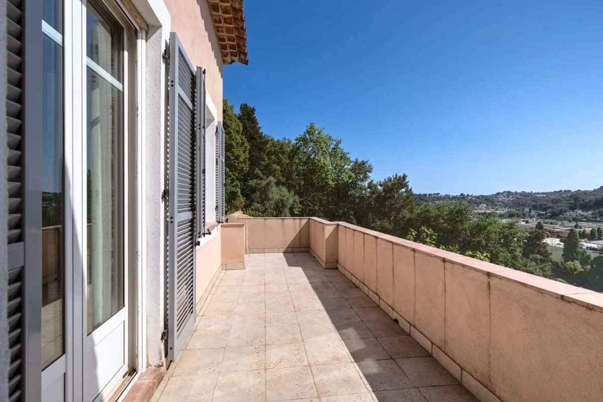 A bright, tiled balcony with peach walls, open shutters, and a clear blue sky overlooking green trees and a distant townscape.