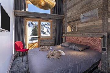 A modern rustic bedroom features wood paneling, a large bed, a red chair, and a balcony offering stunning snowy mountain views.