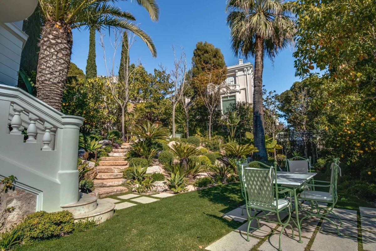 A lush, terraced garden with palm trees, stone stairs, and a light green outdoor dining set. A grand white house is visible under a blue sky.