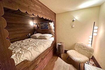 Charming chalet-style bedroom with a carved wooden alcove bed, white duvet, furry chair, and modern multi-bulb lamp.