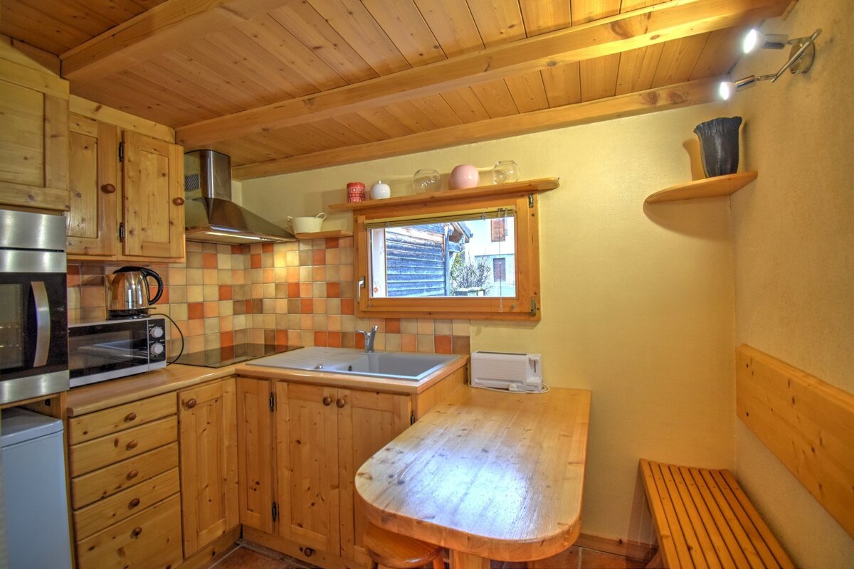 A cozy, compact kitchen with light wood cabinets, ceiling, and a built-in dining table and bench. It features a tiled backsplash and appliances.