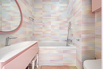 A cheerful bathroom with walls of iridescent pastel tiles in pink, yellow, and blue. Features a pink vanity, white sink, bathtub, and a round mirror.