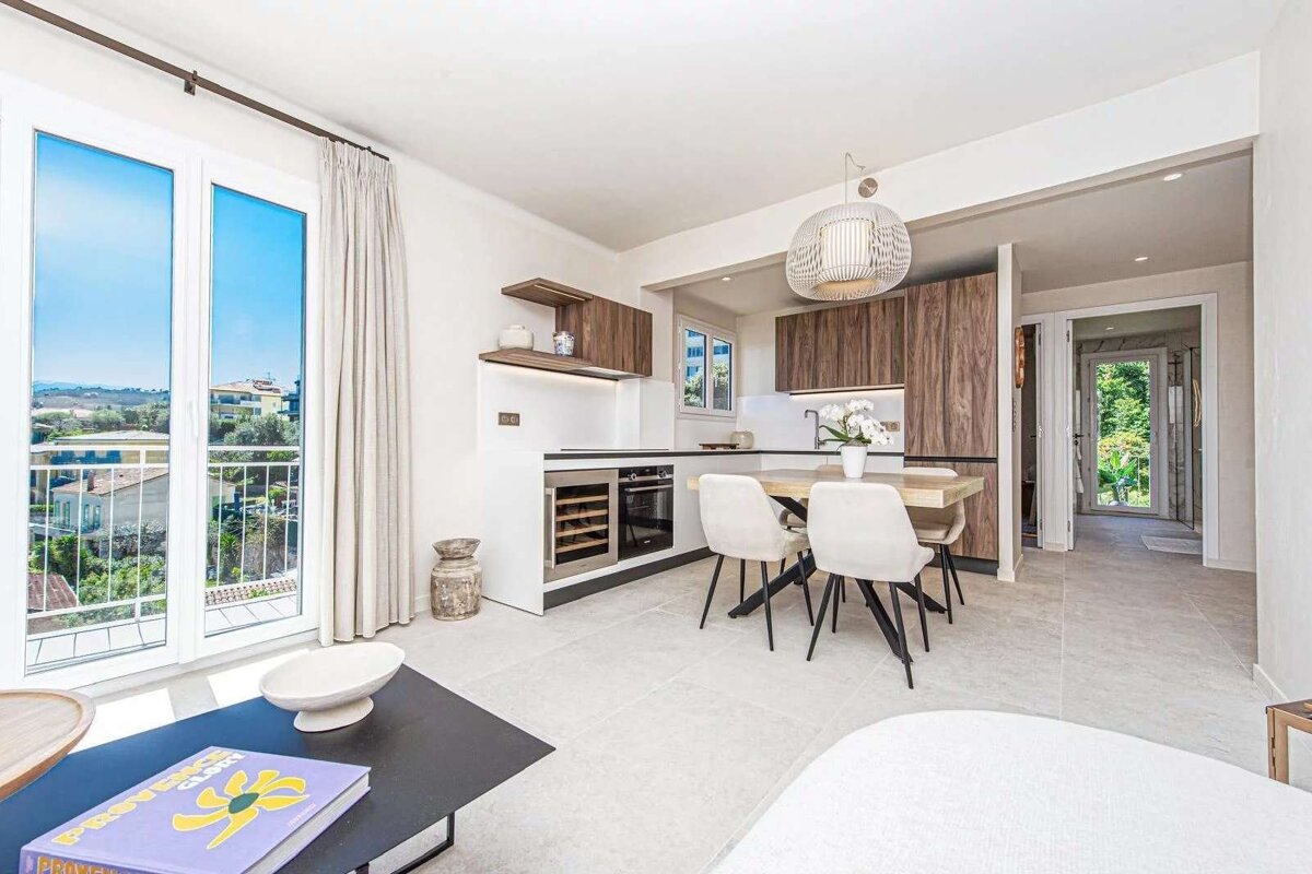 Bright, modern open-plan kitchen and dining area with a large window revealing a scenic view of houses and trees. Sleek cabinets and a stylish dining set.