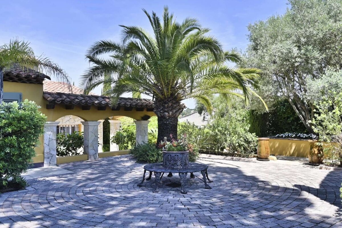 A sunny Mediterranean courtyard features a large palm tree, a circular paver driveway, an arched building, and lush greenery under a clear sky.