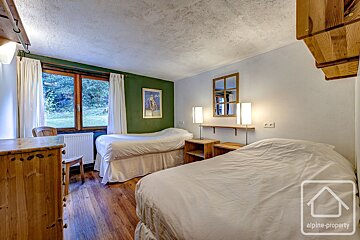 Rustic bedroom with two single beds, a large window overlooking greenery, and a mix of white and dark green walls. Features wooden furniture and floor.