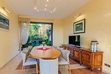 A warm dining area with a wooden table, light yellow walls, and a large glass door overlooking a lush garden. A TV and wooden sideboard are also visible.