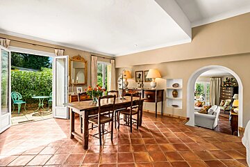 A cozy dining room with terracotta tiles, wooden furniture, open doors to a garden patio, and an arched doorway to a living room.
