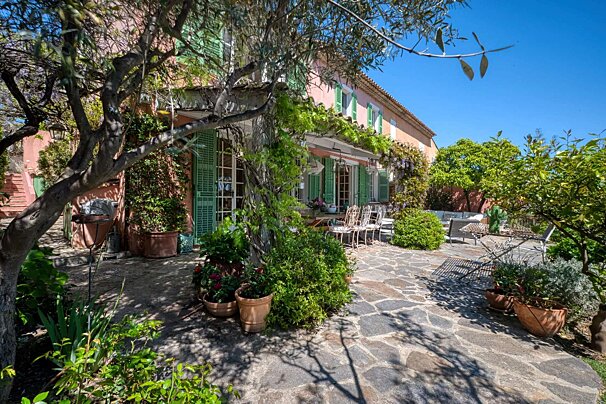 A pink house with green shutters is surrounded by potted plants