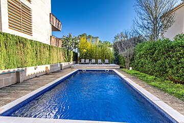 A bright blue swimming pool with loungers on a deck, surrounded by green hedges and a modern building under a clear blue sky.