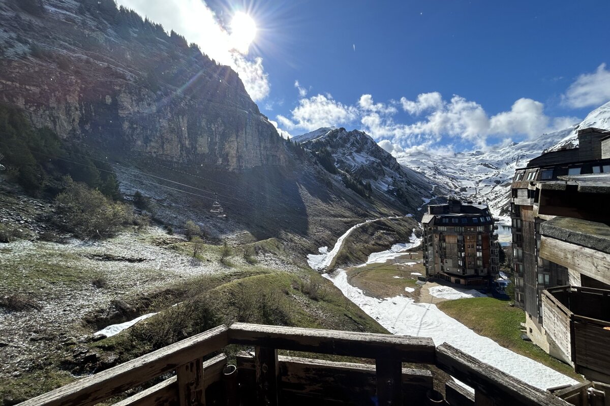 View of a sunlit mountain valley with melting snow, ski resorts, and a wooden balcony in the foreground.