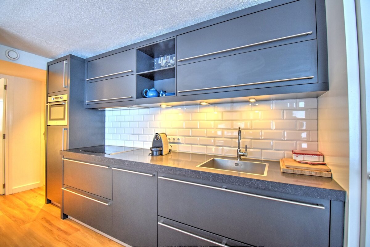 Sleek modern kitchen with dark grey cabinetry, white subway tile backsplash, bright under-cabinet lighting, a sink, and a coffee maker.