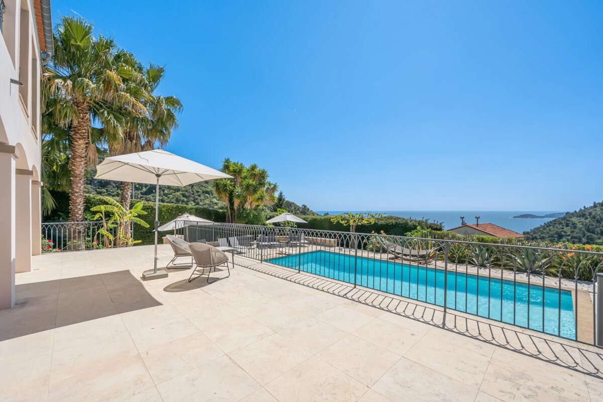 Sunny poolside patio with palm trees, lounge chairs, and a stunning ocean view under a clear blue sky.