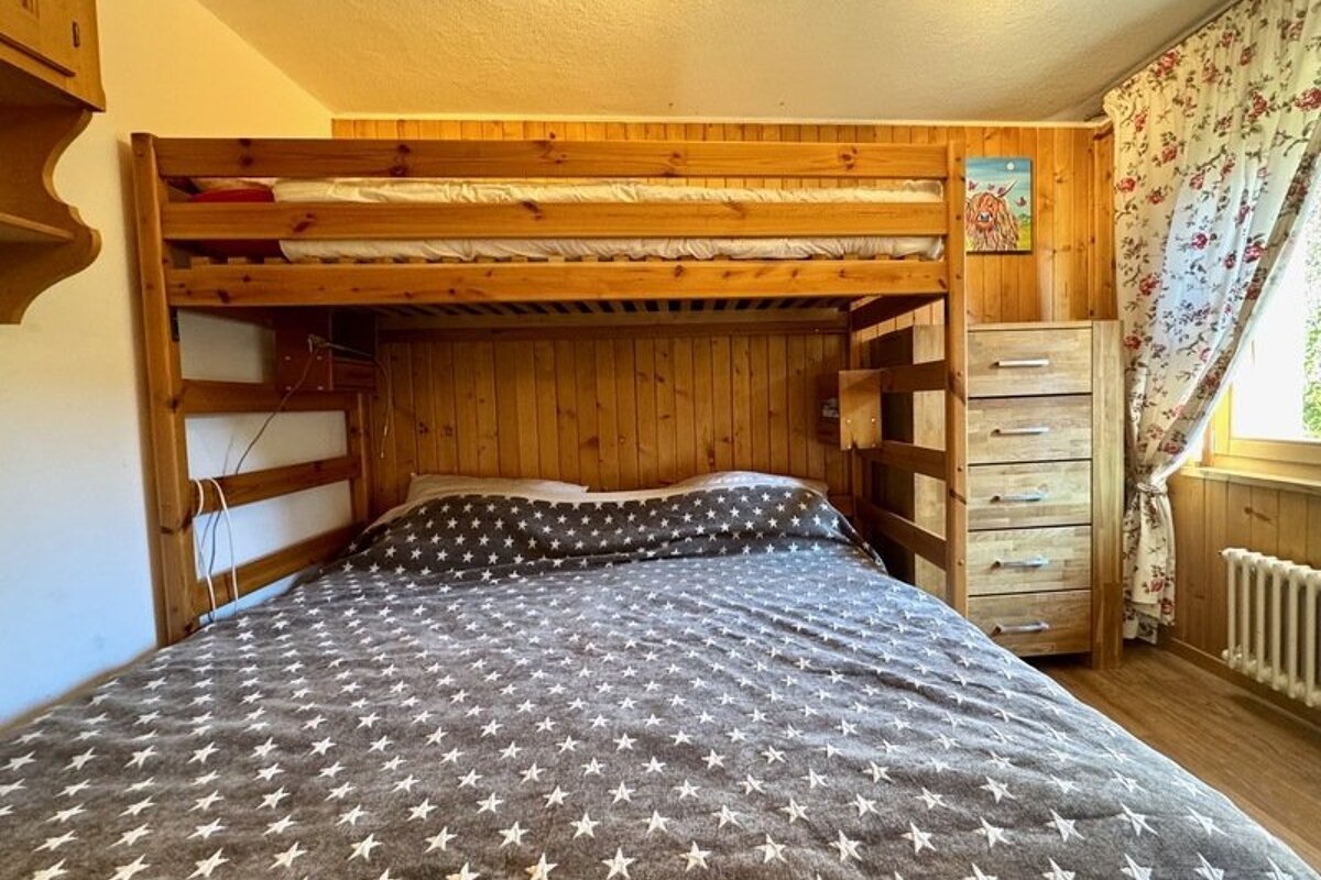 Cozy wood-paneled bedroom with a large wooden bunk bed featuring a grey star-patterned duvet, a dresser, and floral curtains.