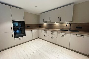 A modern kitchen with white cabinets, dark wood countertops/backsplash, black appliances & handles, and light tiled floors. Under-cabinet lighting illuminates the space.