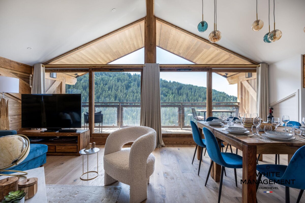A luxurious chalet interior blends rustic and modern design, featuring living and dining areas with large windows that offer stunning mountain forest views.