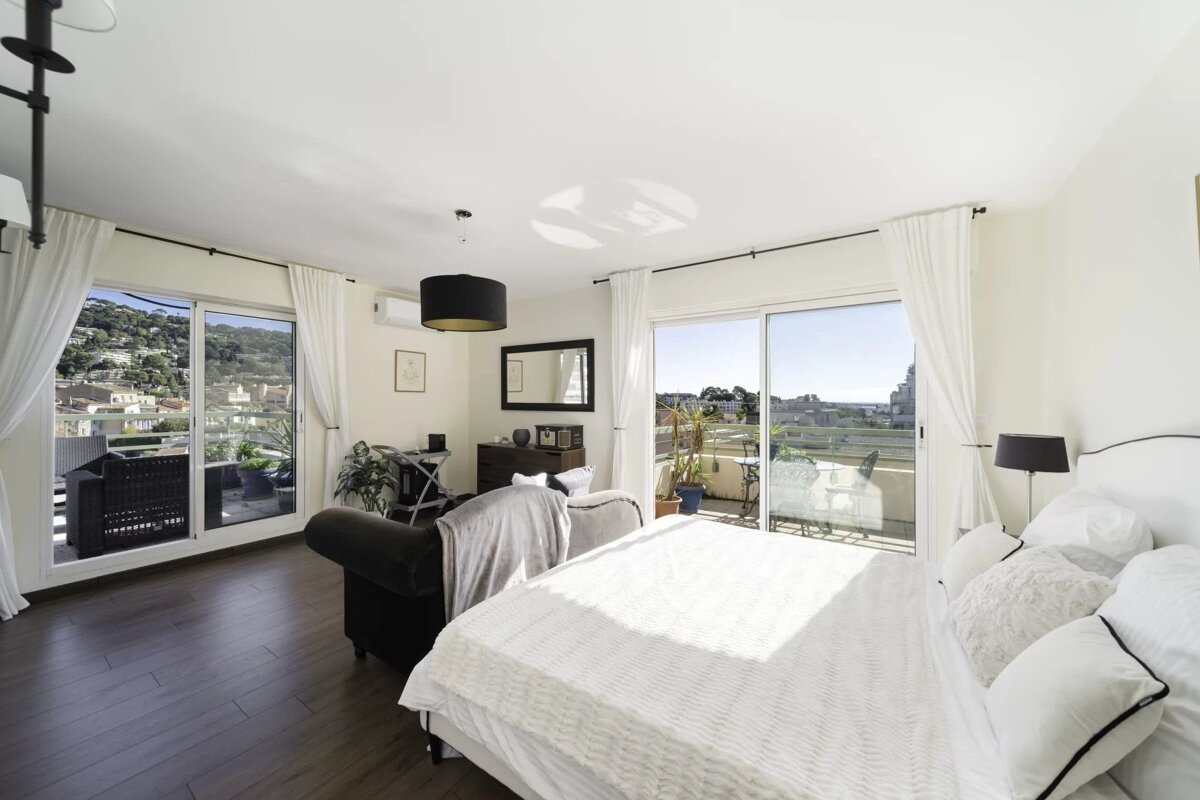 Bright, modern room with a bed, sofa, dark wood floor, and large glass doors leading to balconies with scenic hill and town views.