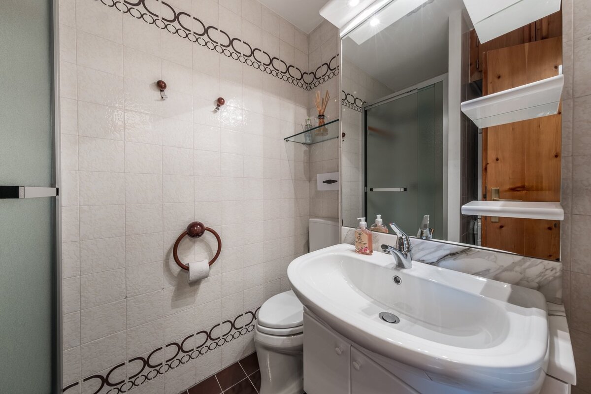 A small bathroom with white tiled walls, brown floor, sink, toilet, and a shower stall. A large mirror and decorative trim are visible.