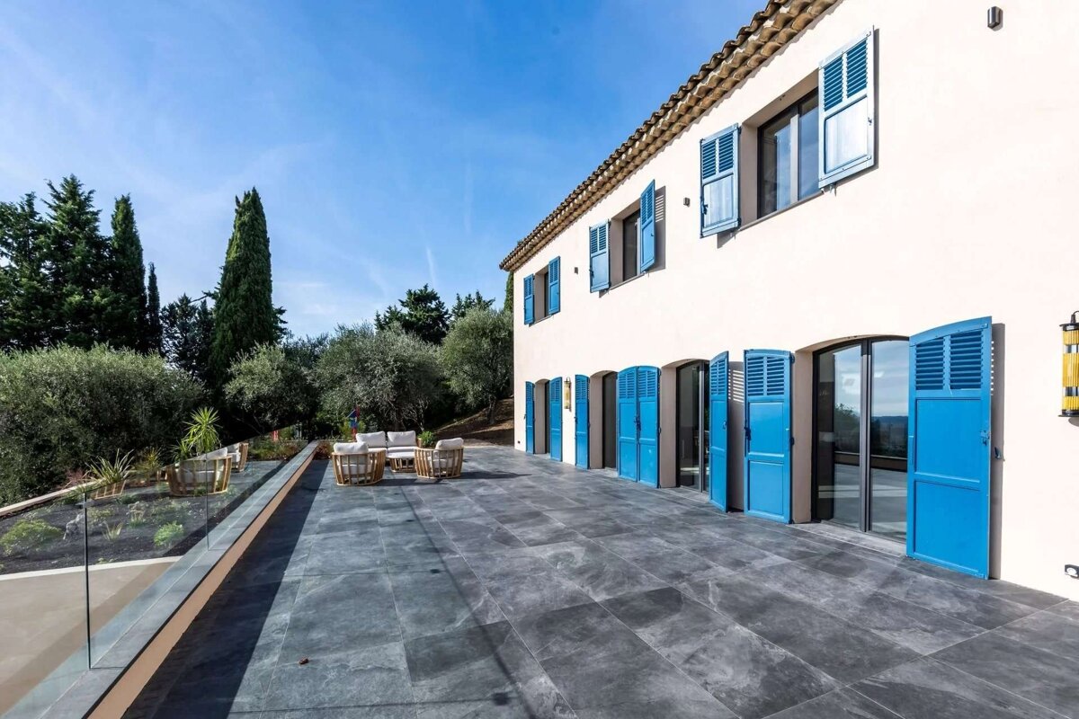 A white house with vibrant blue shutters stands beside a large gray paved patio featuring outdoor seating. Lush green trees and a clear blue sky complete the scene.