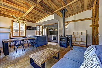 A rustic-modern open-plan room with a dark kitchen, wooden dining table, blue sofa, wood-burning stove, and a prominent wood-planked ceiling.