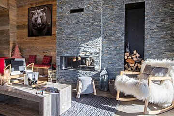 A chic chalet living room with wood walls, a modern stone fireplace, bear art, plush seating, and neatly stacked firewood, bathed in sunlight.