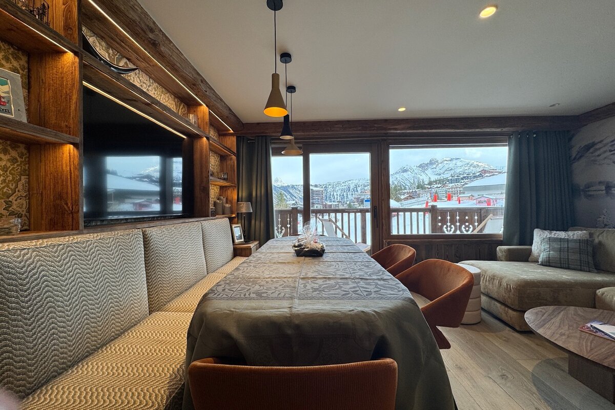 Modern chalet dining room with a long table, textured banquette, and large windows overlooking a snowy alpine resort.