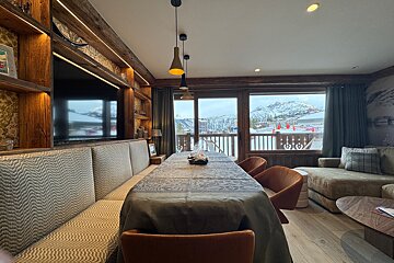 Modern chalet dining room with a long table, textured banquette, and large windows overlooking a snowy alpine resort.