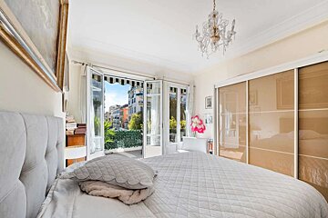 A bright bedroom with a quilted white bed facing open French doors leading to a balcony with city views and lush greenery, under a chandelier.